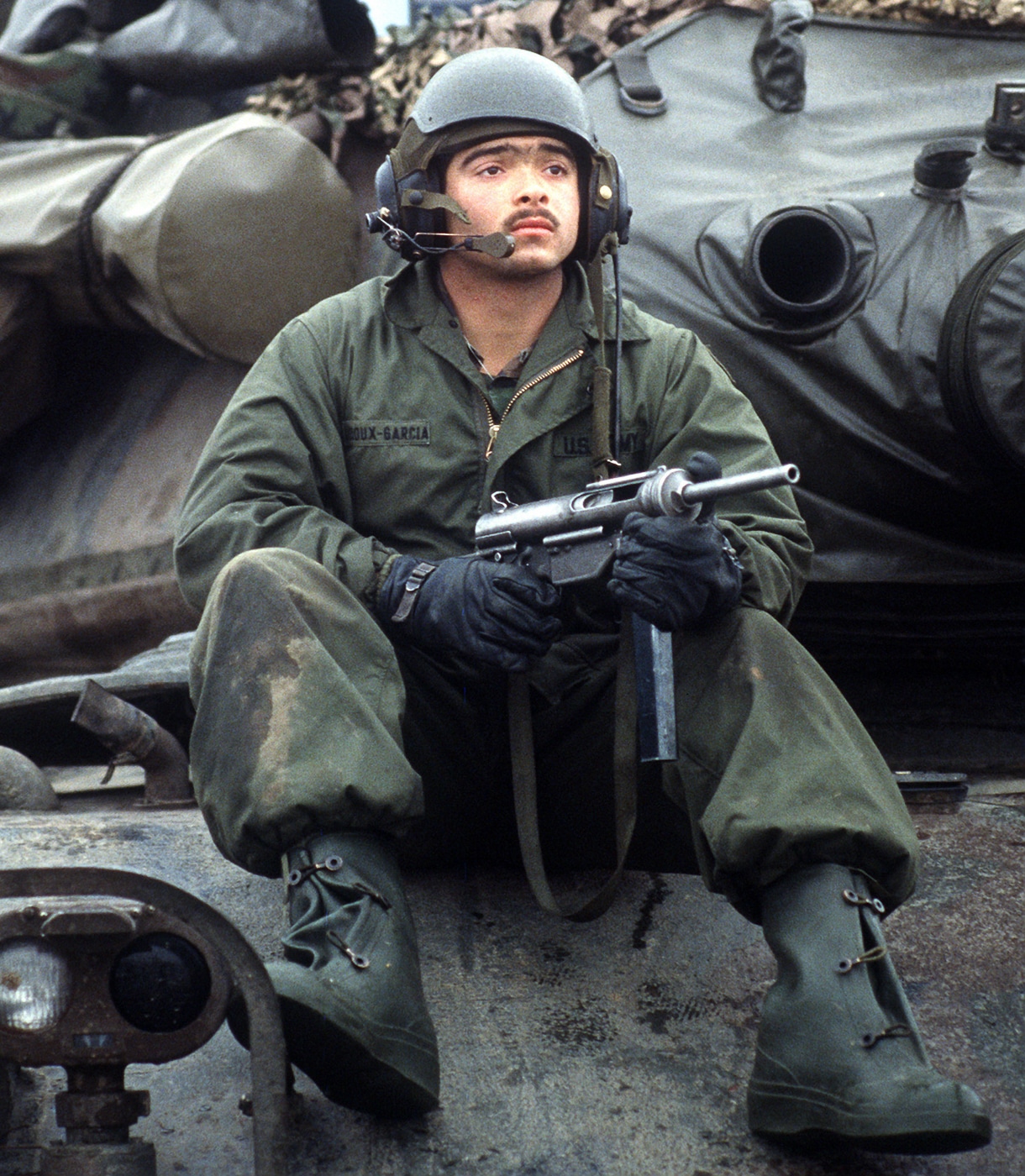 An American soldier in combat gear holding an M3A1 Grease Gun submachine gun during REFORGER 85 military exercises in West Germany with other troops visible in the background. Open bolt firearms remained military standard. Closed bolt guns replaced them gradually. M3A1 Grease Gun used open bolt operation. American submachine guns cost little to produce. Open bolt designs enabled mass production. Military weapons prioritized reliability over precision. Submachine gun service lasted decades. Open bolt mechanisms proved extremely durable.