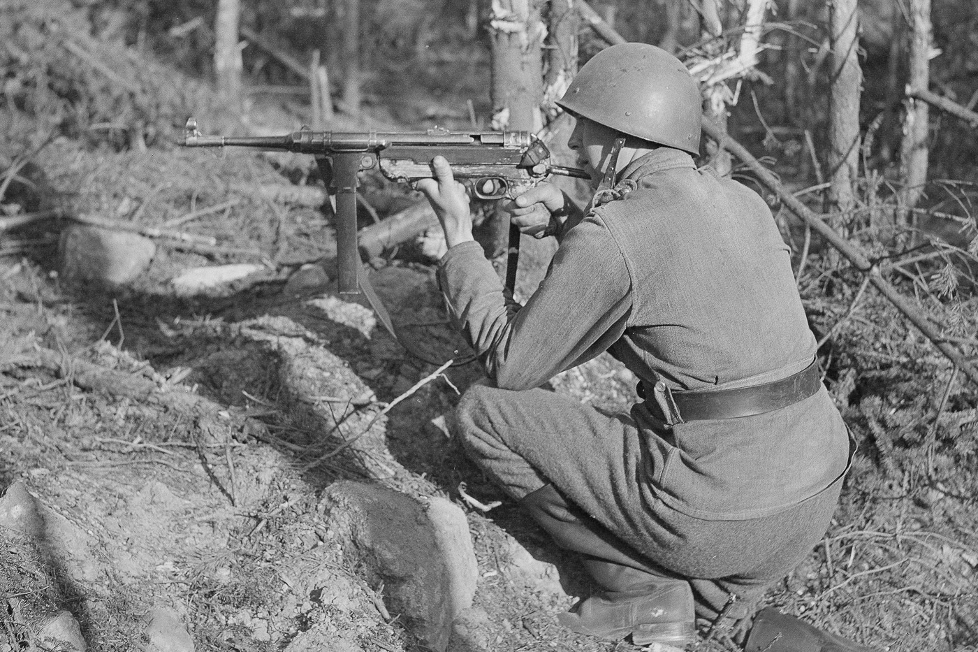 A Finnish military serviceman engaging enemy forces from a crater position while operating a German MP-40 submachine gun showing the practical combat application of open bolt weapon systems. Open bolt versus closed bolt affects battlefield reliability. MP-40 employed proven open bolt operation. Closed bolt weapons require more protection. Finnish forces trusted open bolt SMGs. Open bolt submachine guns perform under stress. Winter warfare demanded dependable firearms. Combat weapons used simple mechanisms. Open bolt designs ensure operational effectiveness.