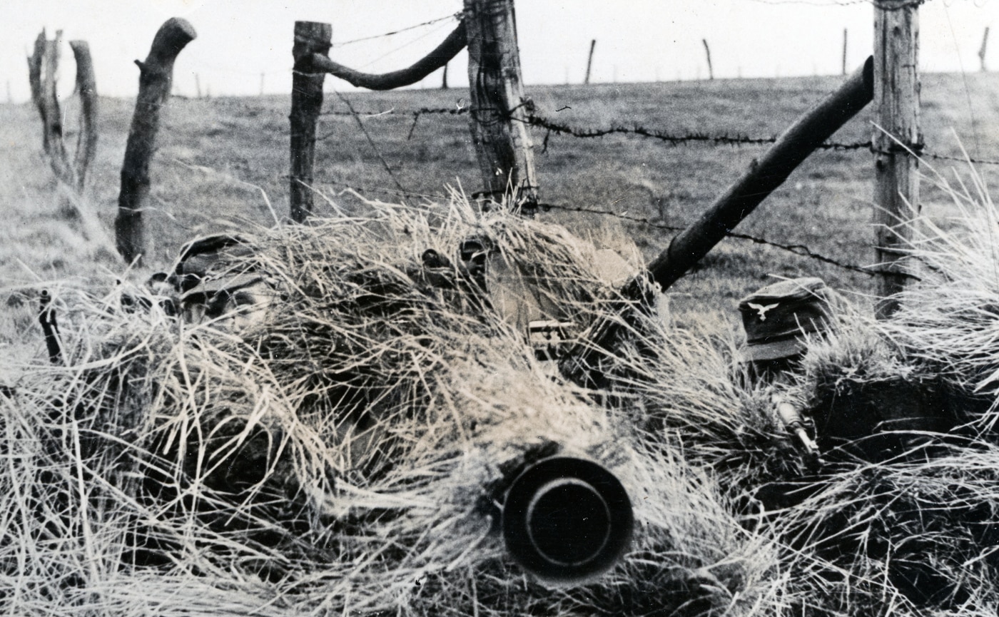 German soldiers crouch behind an 8.8cm Raketenwerfer 43 Püppchen anti-tank rocket launcher that has been concealed within a hay pile to create an ambush position. Wehrmacht crews camouflaged their rocket launchers carefully. The Püppchen required good concealment to survive. German anti-tank teams set up ambush positions. Raketenwerfer 43 gunners needed cover from detection. The hay pile hid the weapon's silhouette. German doctrine emphasized camouflaged firing positions. Anti-tank crews waited for Allied armor. The Püppchen worked best from hidden locations.