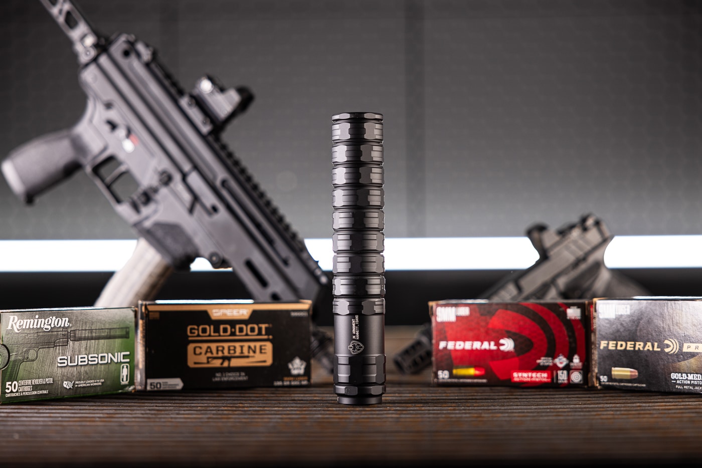 An overhead view displaying the JK Armament 155 RPX 9mm suppressor with accompanying pistols and ammunition boxes spread across a surface for evaluation purposes. The JK Armament 155 RPX 9mm suppressor review examines real-world performance. Handgun silencers soften recoil impulse noticeably. Federal and Remington ammo provided test consistency. The suppressor attaches via HUB mounting interface. Lightweight construction benefits extended shooting sessions. Sound suppressors enhance range communication. Aluminum baffles keep overall weight minimal. The RPX 9mm earns praise for versatility.