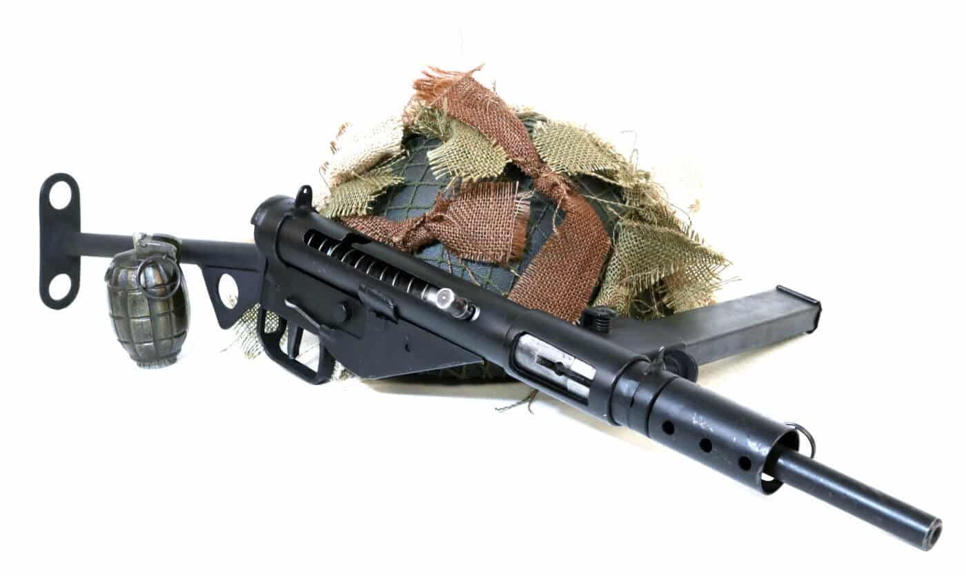 A World War II British Sten submachine gun displaying its crude stamped metal construction, side-mounted magazine, and bare-bones open bolt firing mechanism against a plain background. Open bolt firearms enabled wartime mass production. Closed bolt guns cost significantly more. Sten SMG exemplified open bolt economy. British submachine guns used minimal materials. Open bolt designs reduced manufacturing expenses. WWII weapons prioritized quantity production. Stamped metal construction lowered costs. Open bolt mechanisms simplified assembly processes.