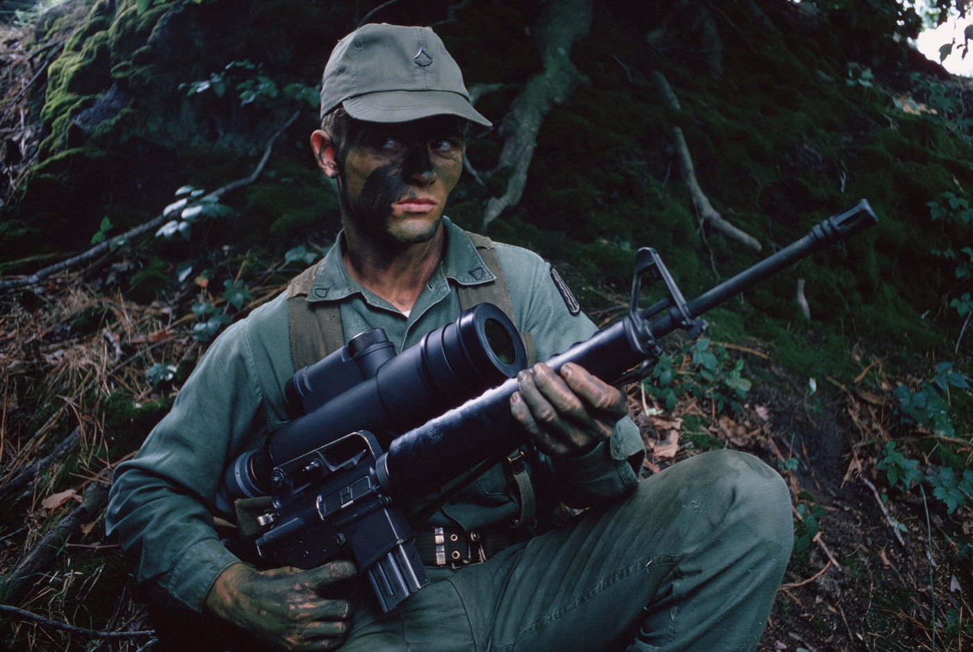 A US soldier in combat fatigues holds an M16A1 rifle with a large cylindrical AN/PVS-2 Starlight scope mounted on top during the Vietnam War era. The Shinenyx G1A review contrasts modern advancements significantly. First-generation night vision amplified existing light sources. Starlight scopes used early image intensifier tubes. Shinenyx night vision gear evolved from military innovations. Vietnam-era devices were heavy and cumbersome. Modern fusion monoculars weigh significantly less. Digital sensors replaced analog tube technology. Thermal imaging complements light amplification now.