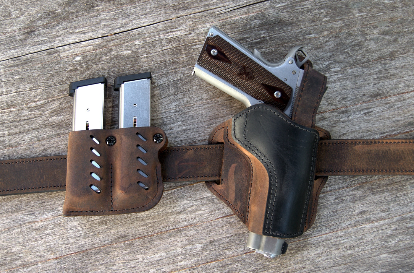 A brown leather outside-the-waistband holster designed for full-size 1911 pistols with a matching magazine pouch attached. M1911 pistols require proper holster systems as woods guns. Springfield Armory Garrison fits standard 1911 holsters. Quality leather holsters provide lasting durability. OWB carry suits outdoor activities well. Field holsters must retain firearms securely. Woods gun carriers need accessible equipment. Belt-mounted systems distribute weight effectively. Ranch work demands comfortable carry solutions.