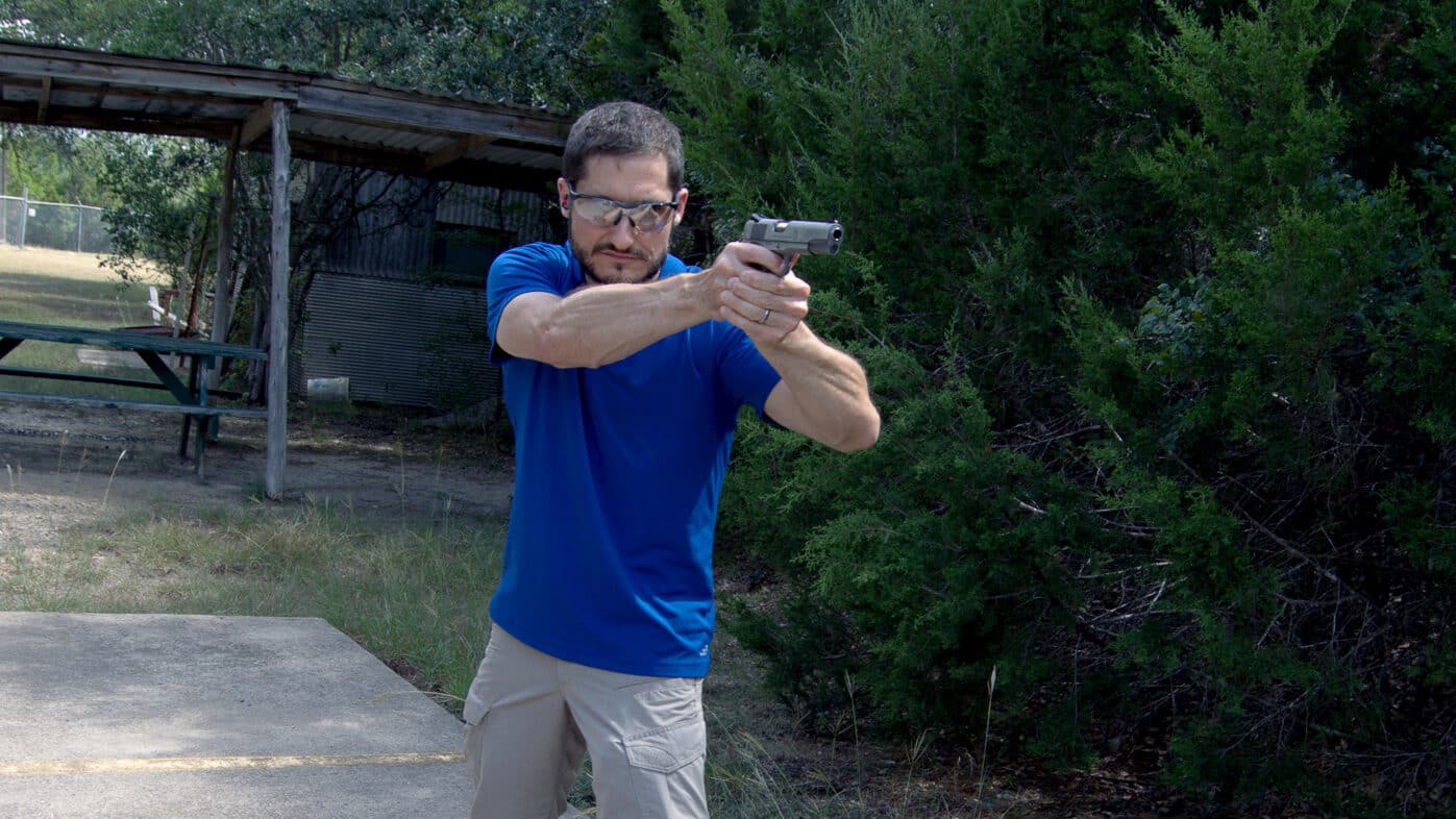 A shooter in outdoor attire firing a stainless steel 1911 pistol at an outdoor range with trees visible in the background. M1911 pistols require practical shooting experience as woods guns. Springfield Armory Garrison handles dynamic shooting scenarios. Live fire training develops defensive skills. Field positions differ from bench rest shooting. Woods gun users practice realistic techniques. The .45 ACP recoil remains manageable. Outdoor ranges simulate hunting conditions. Practical marksmanship builds shooter confidence.