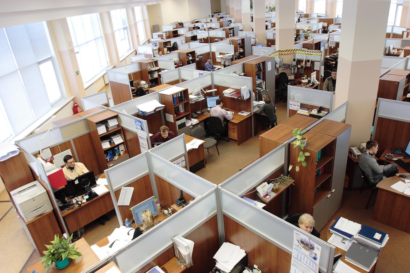 A busy open-plan office shows numerous workstations with employees at desks, limited physical barriers, and close quarters between workers. Concealed carry at work involves crowd awareness. Workplace environments contain innocent bystanders. Office CCW demands situational awareness. CCW at the office requires responsible decision-making. Crowded workplaces increase defensive challenges. Professional spaces need careful consideration. Concealed carry at work means protecting others. Office defense involves understanding surroundings.