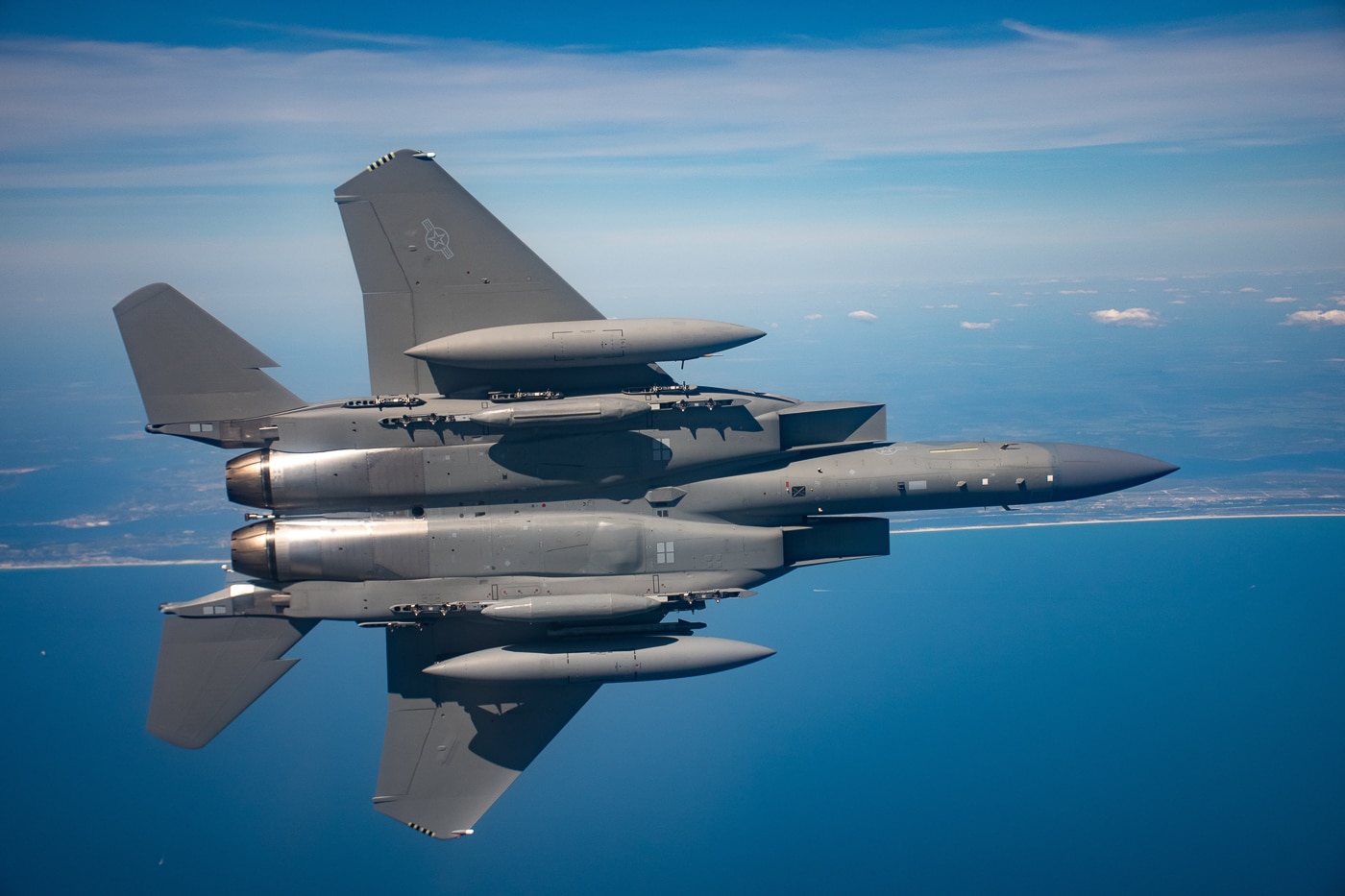 The underside of an F-15EX Eagle II is visible during its first test flight from Eglin Air Force Base, revealing the fighter's weapons pylons, conformal fuel tanks, and twin engine nacelles from below. The belly view shows hardpoint mounting locations. The Eagle II carries weapons on multiple stations. Conformal fuel tanks attach flush to the fuselage. Twin General Electric engines power the fighter overhead. The first test flight validated aircraft systems. Eglin AFB launched the inaugural F-15EX mission. The underside reveals the deep magazine capacity. Boeing engineers monitored the historic flight.