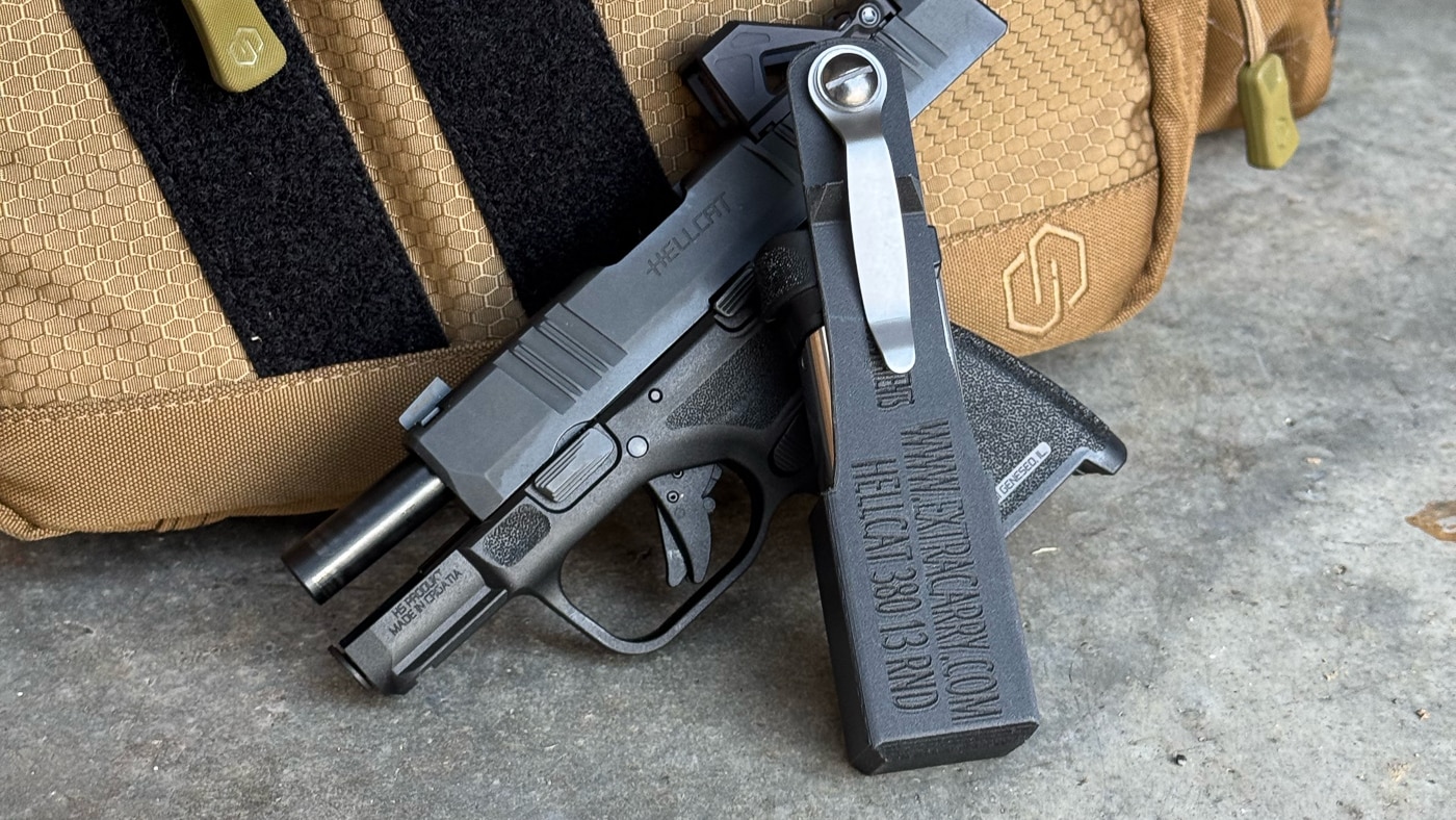 The ExtraCarry magazine pouch is displayed among everyday carry items including a Springfield Hellcat pistol, pocket knife, flashlight, and other EDC essentials, illustrating how the carrier integrates into a complete concealed carry system. ExtraCarry reviews showcase system integration. Magazine carriers complement EDC loadouts. Concealment gear coordinates with defensive equipment.