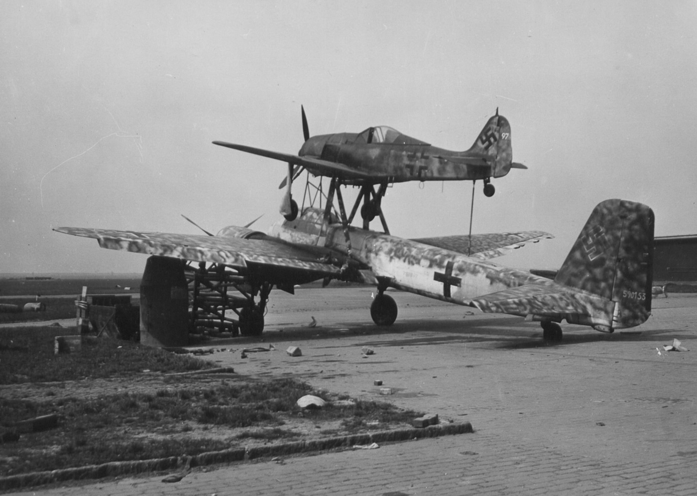 A composite Mistel weapon system consisting of a Focke-Wulf Fw 190 fighter mounted atop an explosive-packed Junkers Ju 88 bomber at captured Merseburg Airport. The Ju 88 bomber served unconventional roles late-war. German composite aircraft used remote guidance systems. Mistel projects transformed obsolete bombers into weapons. Fw 190 pilots controlled unmanned lower aircraft. Desperate measures characterized final war years. Explosive payloads replaced normal bomb loads. American forces discovered experimental German equipment. Captured systems revealed advanced weaponry concepts.