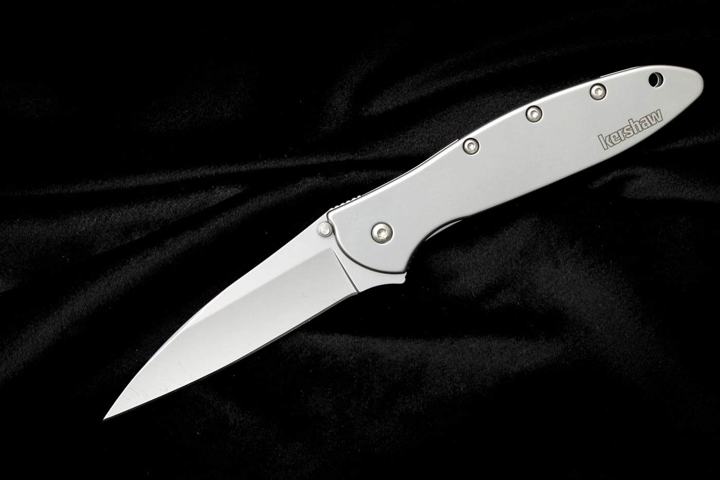 A Kershaw Leek folding knife is displayed showing its distinctive slim profile, stainless steel handle, and assisted-opening flipper mechanism designed by Ken Onion. Kershaw knives popularized assisted-opening technology. The Leek weighs only three ounces. Affordable knives feature 14C28N Swedish steel. Ken Onion designed revolutionary folding mechanisms. Hollow-ground blades create thin cutting edges. Kershaw models include manual safety locks. The flipper tab provides swift deployment. Affordable knives offer radiused handle construction. Three-inch blades balance portability with utility.