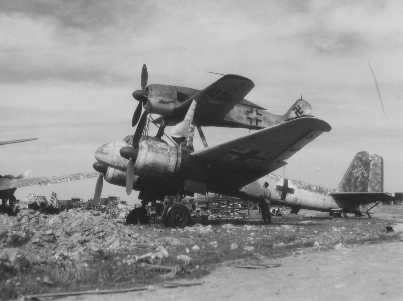 An American soldier from the 439th Troop Carrier inspecting a captured Mistel composite aircraft at a French airbase on May 4, 1945. The Ju 88 bomber participated in experimental weapon programs. Allied troops examined German aviation innovations. Composite aircraft combined fighter and bomber airframes. French airbases yielded abandoned Luftwaffe equipment. War's end revealed secret German projects. American units documented captured enemy technology. Piggyback configurations enabled remote guidance attacks. Historical photographs record liberation discoveries.
