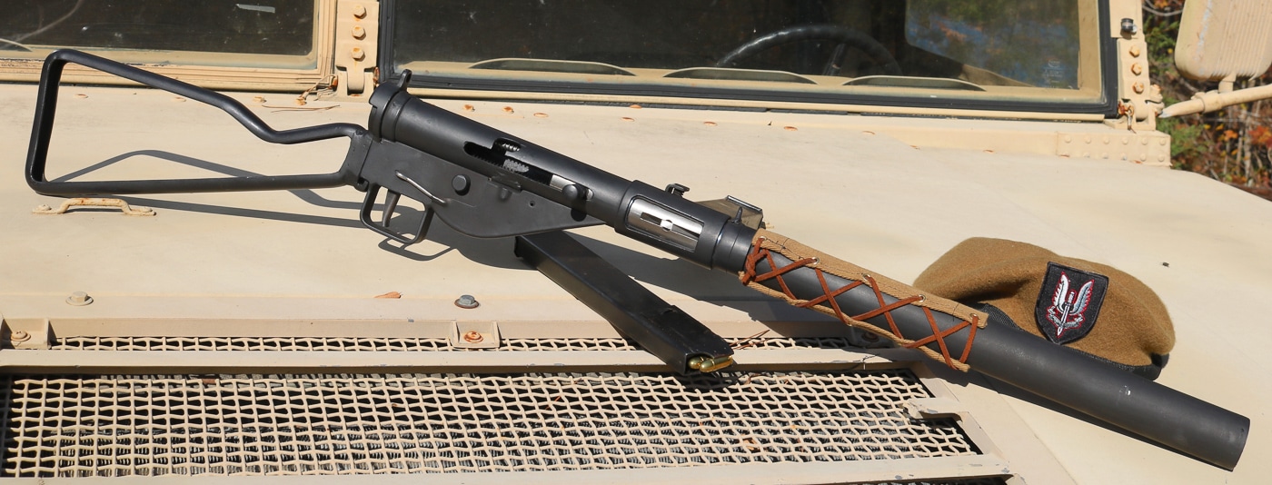 A Sten Mk IIS submachine gun resting on the hood or fender of a military vehicle, showing the weapon in a tactical field context with its distinctive canvas-covered suppressor and side-mounted magazine clearly visible. The suppressed Sten Mk IIS served combat operations. Special forces deployed silenced weapons tactically. The 9mm SMG fired subsonic ammunition. British commandos utilized sound-suppressed firearms. Wire-mesh baffles reduced muzzle noise. Covert missions required stealthy armament. Elite operators valued silenced SMGs. Field conditions tested suppressor effectiveness.