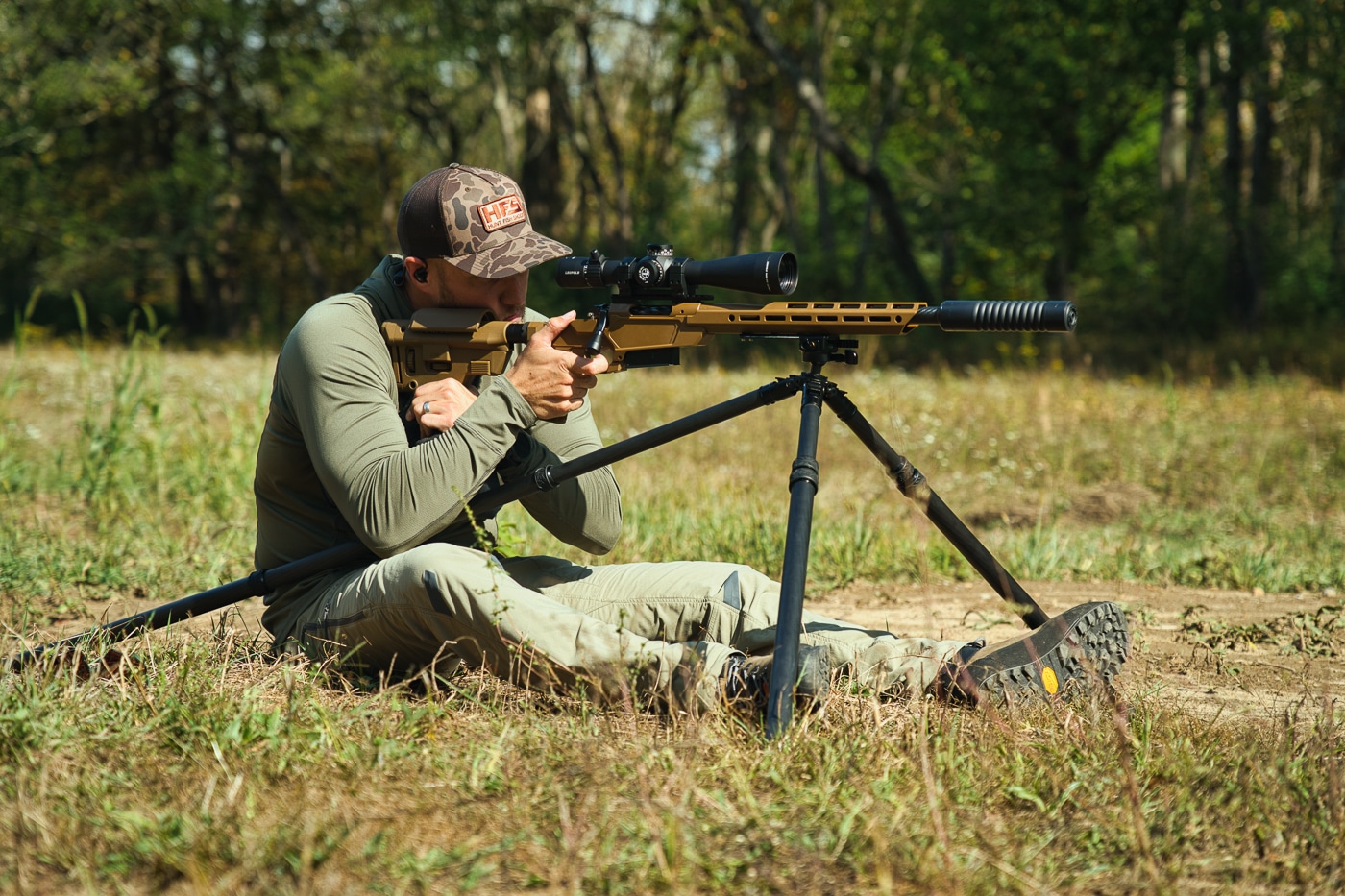 A shooter sits on the ground at a shooting range with a scoped rifle mounted on a Tricer-HK tripod with the legs independently adjusted for stability. The Tricer-HK tripod review confirms versatile positioning capability. Ultra light Tricer-HK tripod evaluation demonstrates seated shooting stability. Adjustable legs accommodate various shooting positions.