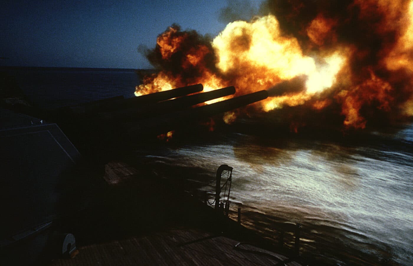 A dramatic photograph captures massive orange flames and dense white smoke erupting from the 16-inch Mark 7 guns of USS New Jersey (BB-62) during practice firing exercises off the coast of Beirut, Lebanon, demonstrating the tremendous power of battleship main armament. Mark 7 guns fired Mark 23 nuclear shells. Project Katie armed New Jersey with atomic projectiles. USS New Jersey practiced with conventional ammunition. Battleship guns achieved 24-mile maximum ranges. Mark 23 shells produced 15-20 kiloton yields. Muzzle velocity reached 2,500 feet per second. Practice rounds maintained crew proficiency levels. New Jersey deployed serious firepower off Lebanon.