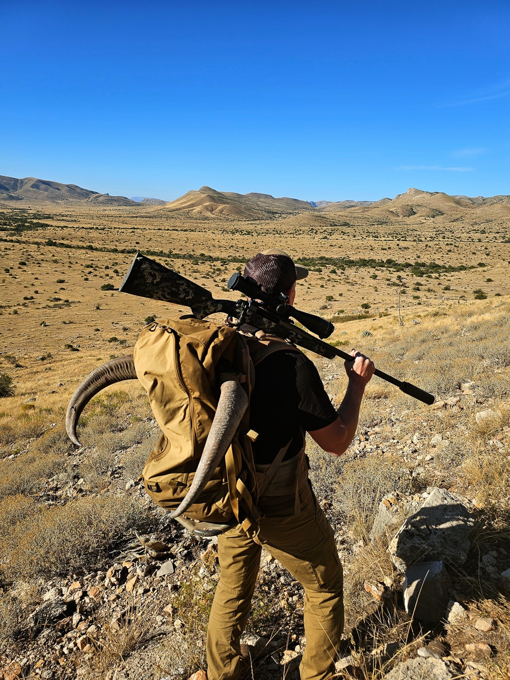A hunter carries a rifle slung over his shoulder while hiking across rugged terrain with mountains visible in the background, illustrating the importance of rifle weight during extended hunts. First hunting rifles should balance weight and durability. Beginner's hunting rifle selection considers carry comfort. Lightweight construction reduces hunter fatigue. Carbon fiber components decrease overall mass. Model 2020 rifles weigh under eight pounds. Extended hikes demand practical equipment choices. Rifle weight affects shot steadiness. Smart hunters prioritize manageable packages.