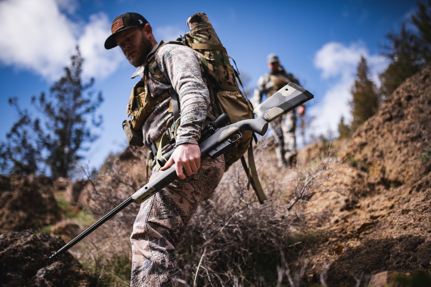 A hunter traverses steep mountainous landscape with a bolt-action rifle strapped across his back, demonstrating the physical demands of backcountry hunting. First hunting rifle weight impacts field performance. Beginner's hunting rifles benefit from modern materials. Unnecessary pounds cause premature exhaustion. Springfield Waypoint rifles emphasize portability. Long-distance hunting requires thoughtful gear selection. 