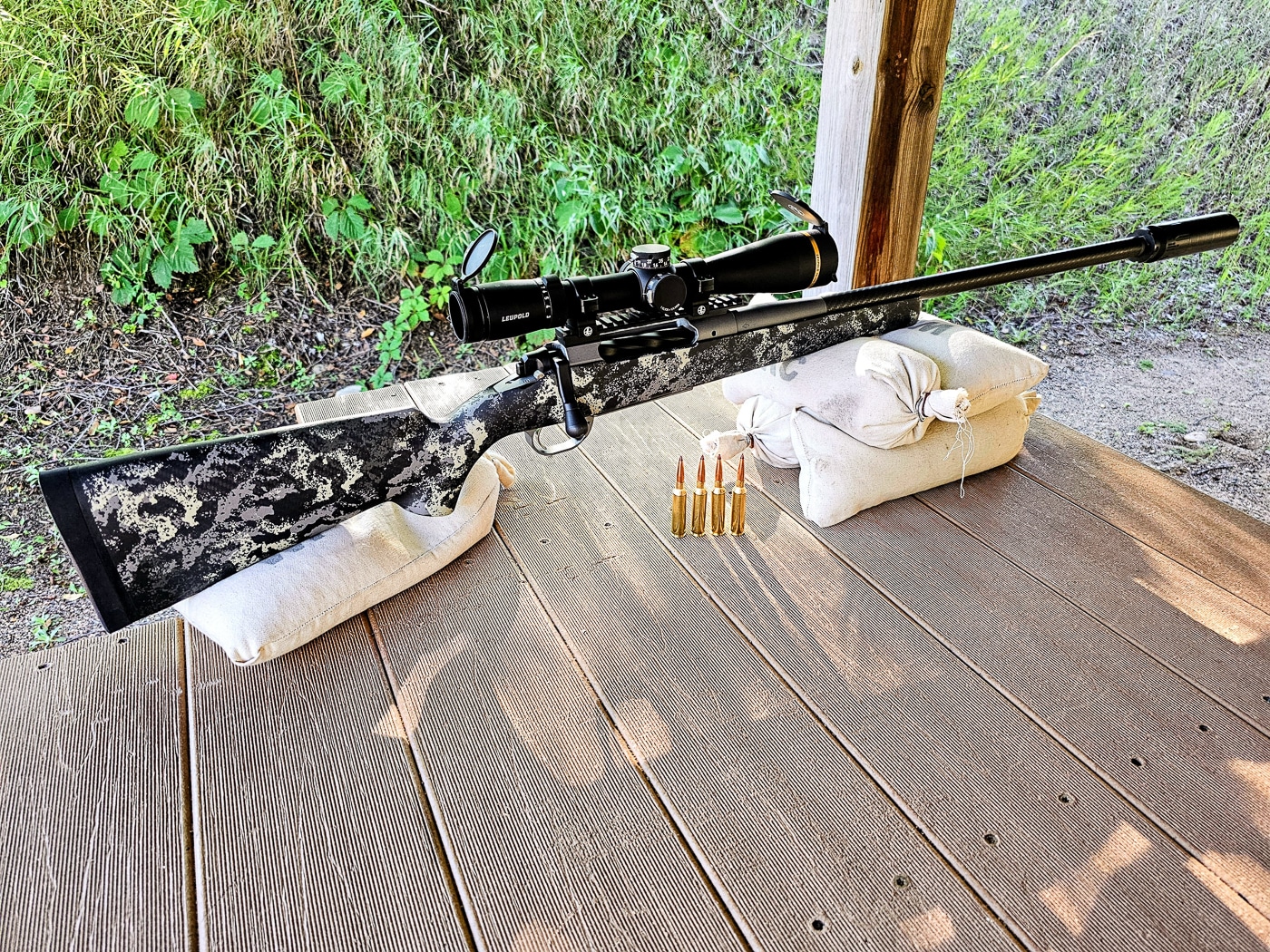 A hunter uses a makeshift sandbag rest to stabilize a bolt-action rifle during an accuracy test at an outdoor shooting range. First hunting rifles require steady shooting platforms. Beginner's hunting rifles shoot accurately from improvised rests. Sandbags reduce rifle movement during firing. Practical shooting positions mirror hunting conditions. Field-ready shooters adapt available equipment. Stable rests improve shot consistency. 