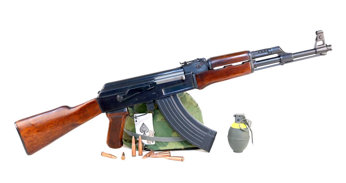 An AK-47 rifle is displayed alongside a military helmet, a grenade, and loose ammunition in a posed arrangement. The AK-47 in Vietnam War combat was considered one of the most effective infantry weapons fielded by any combatant. Kalashnikov rifles gave Viet Cong fighters reliable firepower in the hot and humid jungle environment. The 7.62x39mm intermediate cartridge fired by the AK-47 delivered significant stopping power at combat ranges. Chinese Type 56 rifles were the most commonly encountered Kalashnikov variant by U.S. troops in Vietnam. AK-47 ammunition was widely available through Soviet and Chinese supply networks supporting North Vietnam. Viet Cong fighters carried grenades and rifles as their standard combat loadout in jungle ambushes. The AK-47 rifle's durability made it ideal for guerrilla warfare in tropical conditions. Vietnam War weapons captured by U.S. forces revealed the sophistication of Communist-bloc small arms development.