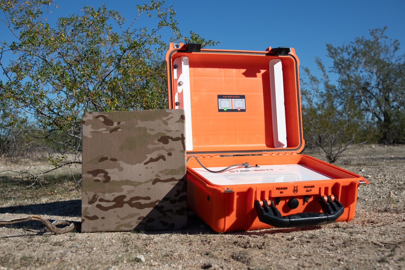 A photo of a StarBatt portable battery kit set up in an outdoor field environment, with cables connecting to satellite communication equipment and rugged terrain visible in the background. Off-grid survival communication tools require dependable power sources. Portable batteries run satellite systems. Base camps need sustained electricity. Overland rigs carry backup power. Starlink Mini units demand consistent voltage. Lithium packs replace fuel generators. Field setups depend on mobile power. Remote cabins benefit from battery kits.