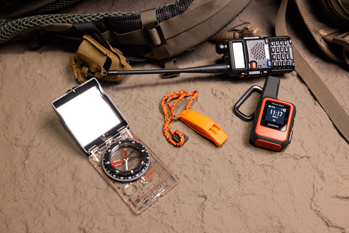 A flat lay photo showing a Baofeng handheld radio, Garmin inReach Mini 2 satellite communicator, an orange emergency whistle, and a compass with a built-in signaling mirror arranged on a neutral surface. Off-grid survival communication tools save lives. Backcountry gear enables emergency contact. Handheld radios allow group coordination. Walkie talkies transmit short-range signals. Satellite messengers reach global networks. The Garmin inReach Mini 2 sends two-way texts. Signal mirrors reflect sunlight for rescue. Emergency whistles produce loud distress tones.