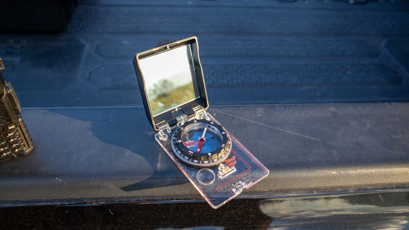 A  photo of a small handheld compass featuring a built-in reflective signaling mirror on its lid, with a directional dial and aiming hole visible for emergency use. Off-grid survival communication tools include signal mirrors. Mirrors reflect sunlight over distances. Compasses orient lost hikers. Reflective surfaces attract aircraft attention. Aiming holes target distant rescuers. Hunters carry combination signaling units. Compact tools serve dual purposes. Backcountry kits demand simple gear.