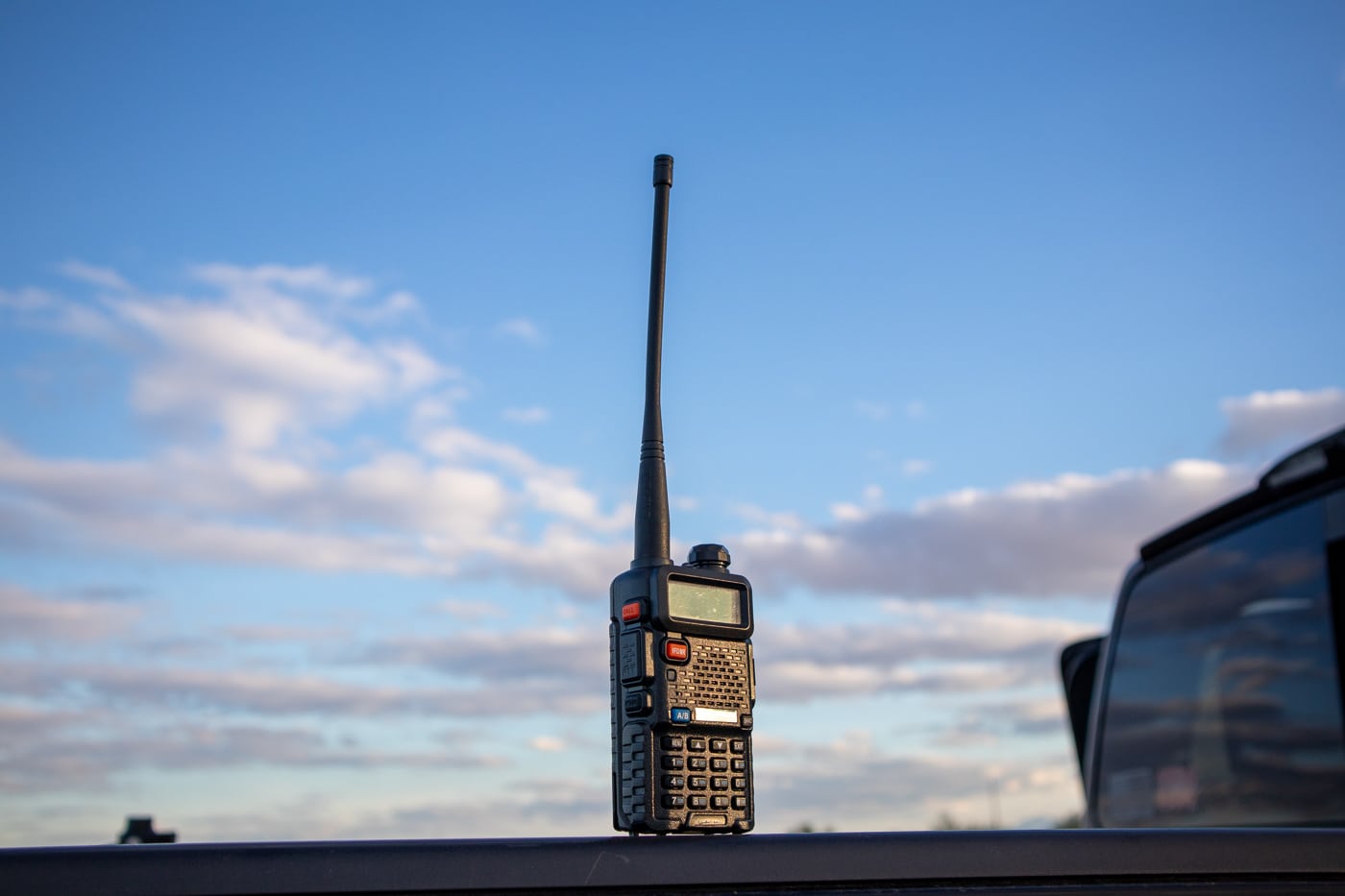A close-up photo of a black Baofeng handheld two-way radio with an extended antenna, LCD display, and push-to-talk button visible on the side. Off-grid survival communication tools include handheld radios. Walkie talkies enable direct contact. Hunters use radios for coordination. Hikers rely on two-way transceivers. Antennas determine signal strength. Terrain affects radio range. Push-to-talk simplifies group communication. Battery life impacts field performance.