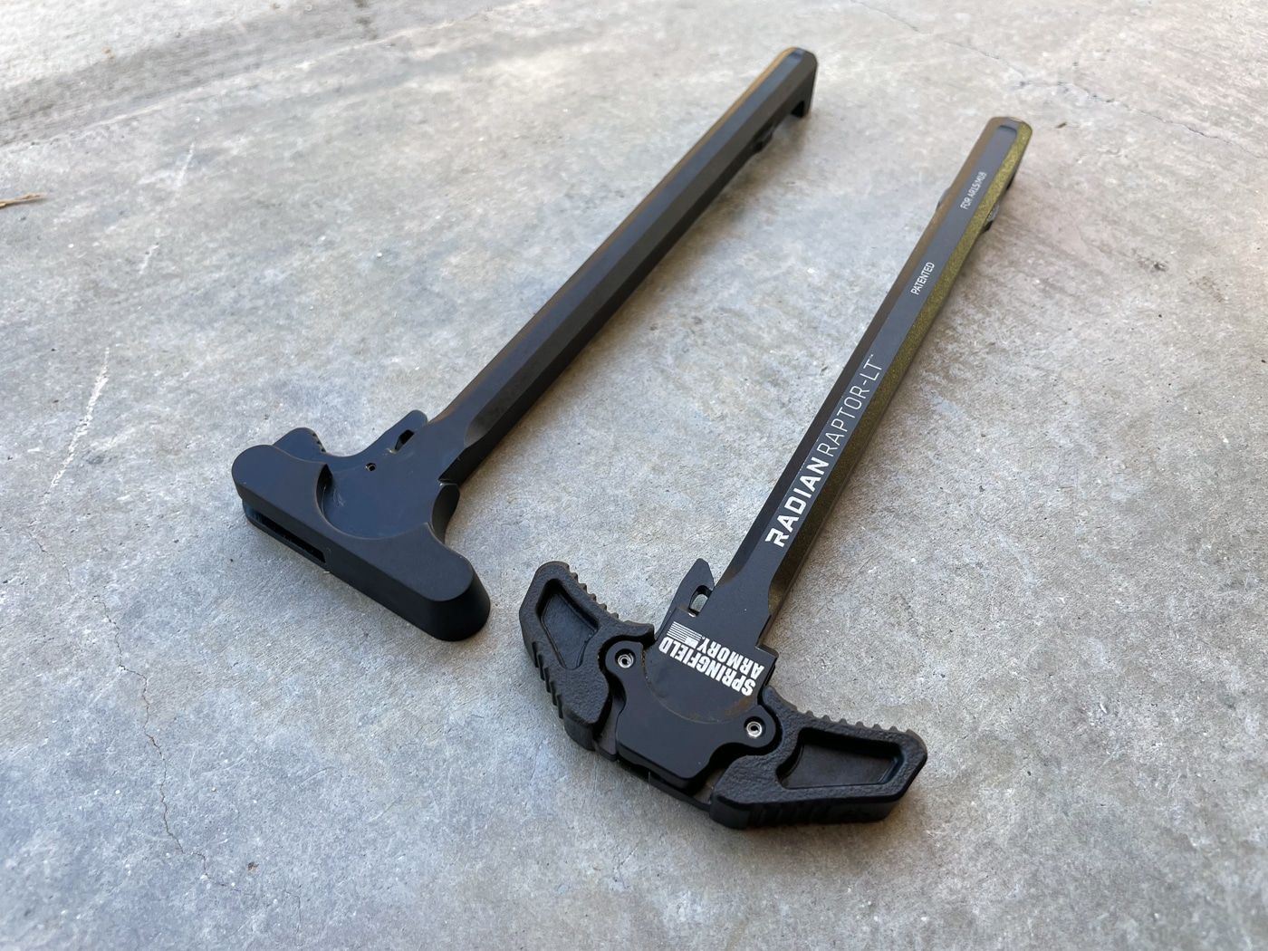 Two AR-15 charging handles are arranged next to each other for comparison, with the smaller rounded mil-spec unit on one side and the larger Radian Raptor-LT with its extended ambidextrous polymer paddles on the other. The Radian Raptor LT review demonstrates size differences. Mil-spec handles measure smaller. Raptor handles extend wider. AR-15 shooters compare options. Oversized levers provide leverage. Standard handles favor right-handers. Ambi handles serve everyone. Upgrades improve manipulation.