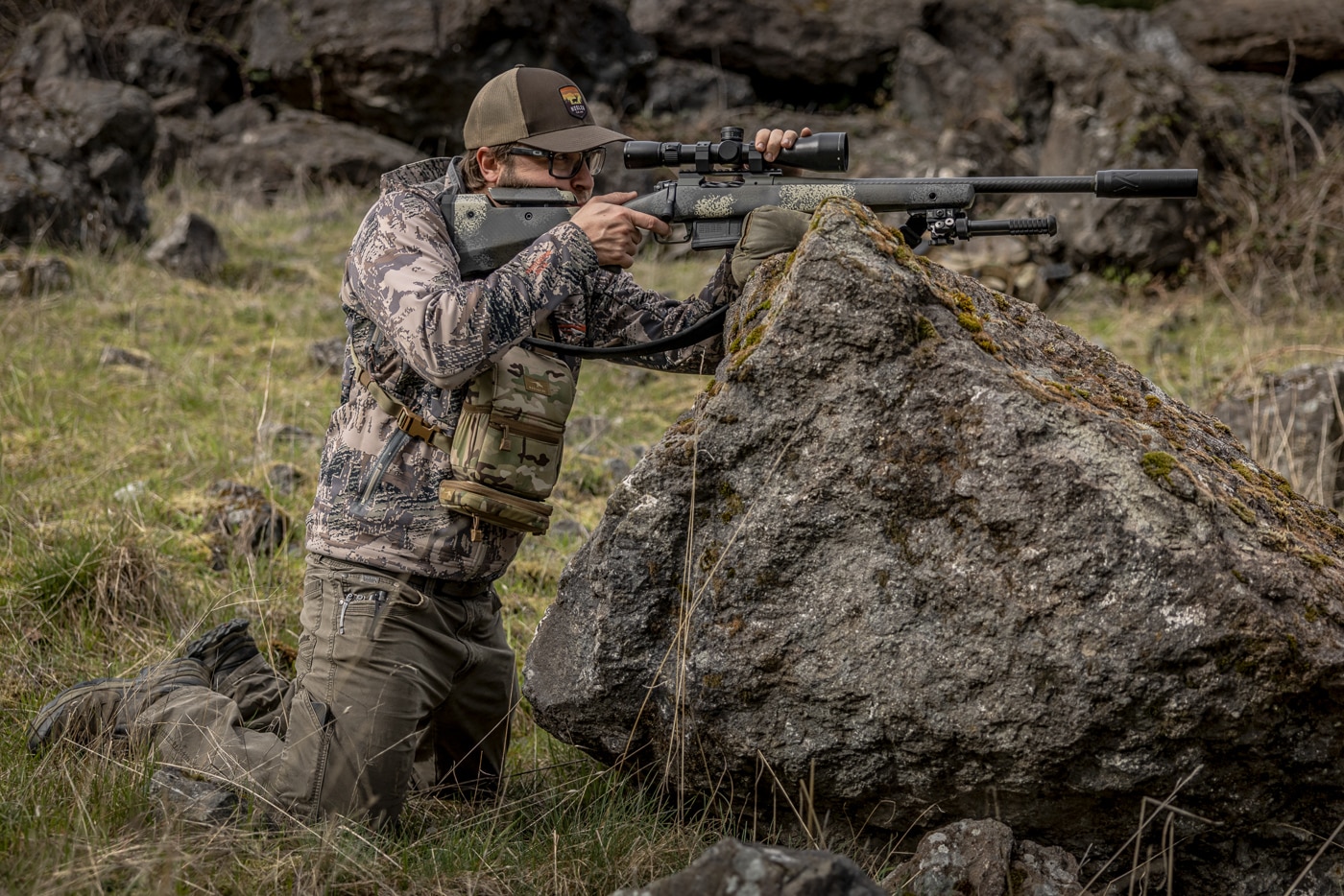 The photo shows the author resting his rifle on a Pint-Sized Game Changer shooting bag from Armageddon Gear, which sits on top of a large boulder. A quality shooting bag is among the most useful tools for building stable hunting shooting positions, since it adapts a rock, stump, or fence post into a steady rest. This particular bag offers a flat side for level surfaces and a notched side that conforms to rounded objects like the boulder pictured. The author prefers the sand fill because it provides more stability than the lighter fill options, even though it adds weight. Bedding the rifle near its center of balance and pressing down on the scope keeps the muzzle from wandering. Whether seated, kneeling, or standing, a shooter can build a solid platform around the bag in moments. It is exactly the kind of versatile gear worth training with before a hunt.