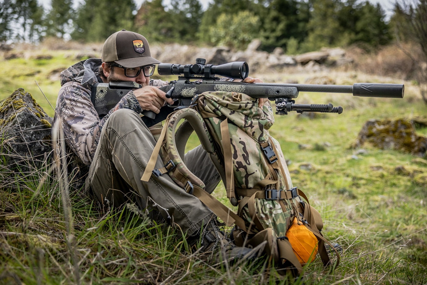 The image shows the author using an EXO MTN Gear K4 frame pack as a rifle rest while shooting in the field. Turning a backpack into rifle support is one of the most weight-conscious hunting shooting positions, since the pack is gear the hunter already carries. The author has used EXO MTN Gear packs for more than 10 years and relies on the K4 system for its comfort and durability on long pack-outs. To build the position, he rests the rifle on the pack frame and pinches the gun to the pack or presses down on the scope. Unlike tripod work, this support benefits from pulling the rifle more firmly into the shoulder to manage recoil. The setup is less stable than a tripod or shooting bag, but it carries no extra weight and works well on elk, deer, and sheep hunts. Putting gear to work this way frees the hunter from hauling sticks or a tripod into the backcountry.