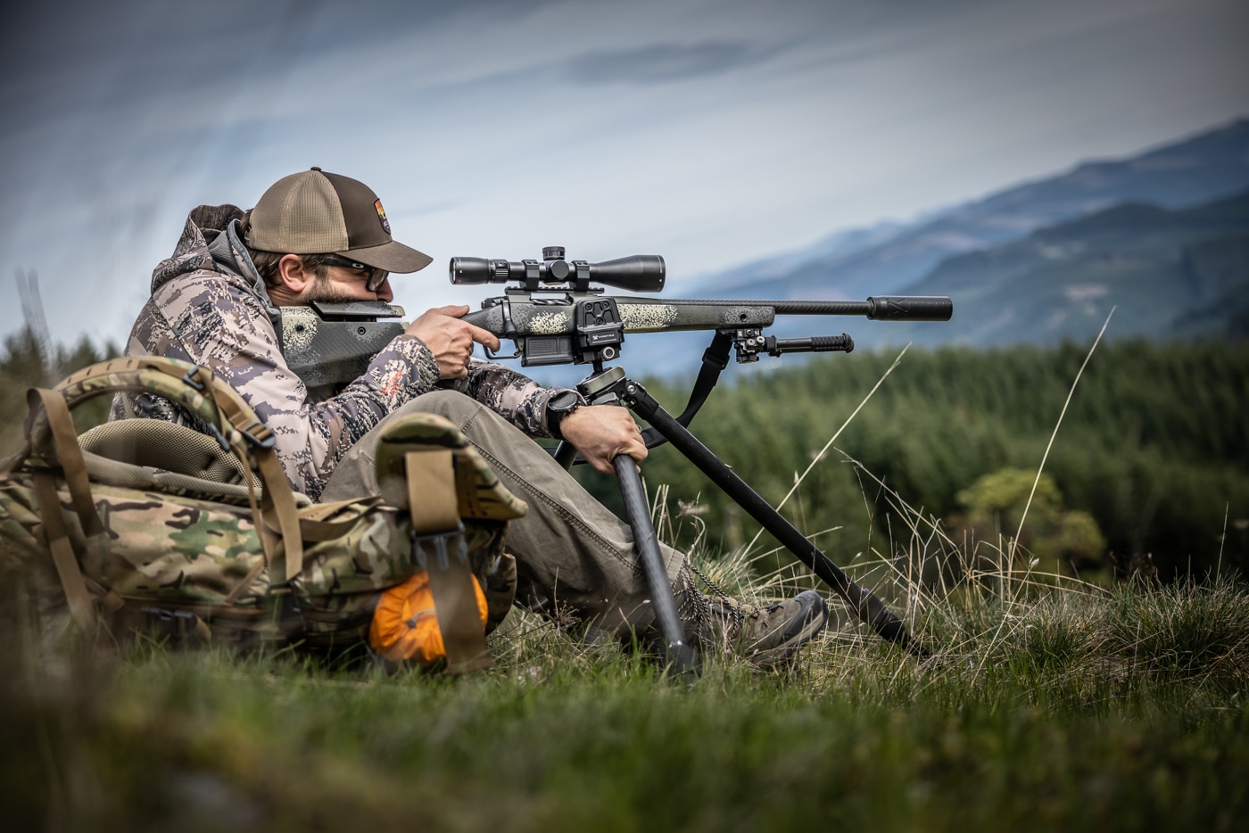 The image shows the author standing in open terrain behind the Vortex Optics Switchback Carbon, a carbon-fiber shooting tripod, with the legs spread for stability. A shooting tripod underpins some of the most reliable hunting shooting positions, from seated work on a slope to standing shots over tall grass. This model balances light weight, a compact folded size, and enough rigidity to steady a rifle for an ethical shot. A single throw lever adjusts the ball head, letting the author level the platform and get on target quickly. Orienting the legs like an arrow pointing toward the target sends recoil straight back for faster follow-up shots. The same tripod also supports a spotting scope or binoculars, which is why versatile gear like this stays in his pack.