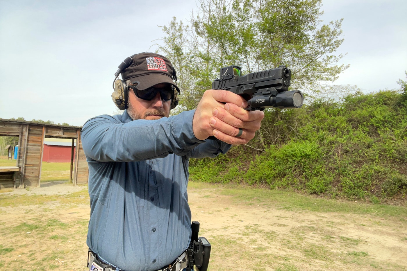 A right-handed shooter draws a pistol from a hip holster and extends it toward a target with a two-handed grip. This photo illustrates the opening movement of the War HOGG Self Eval, a performance-on-demand drill that tests marksmanship and gun handling with no warm-up. The draw, or pistol presentation, is the first timed event in the drill and accounts for the draw-to-first-shot measurement. Running the War HOGG drill cold gives a shooter an honest read on real shooting performance under the clock. The skill builder is meant to be shot at the start of the month, recorded, and retested later to track improvement. A smooth, repeatable draw stroke is one of the gun handling skills this self-evaluation exercise is built to expose and refine.