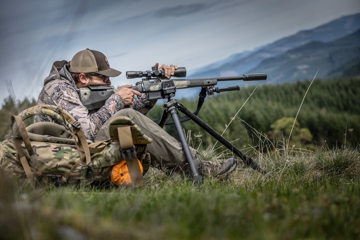 A hunter sits behind a tripod-mounted rifle in the field, his firing-side knee up and his elbow braced against his thigh. Among the hunting shooting positions covered for the field, tripod sitting may be the most useful because it sets up fast and holds steady on varied terrain. The low seated posture drops the shooter's center of gravity and shrinks the wobble zone behind the rifle. Mounting the gun just ahead of the magwell keeps it balanced on the head and makes small aiming corrections easy. Pushing firmly but not hard on top of the scope helps the author control recoil and keep the sight picture settled. For a hunter who has only ever shot from a bench, learning this position is a major step toward field-ready accuracy.