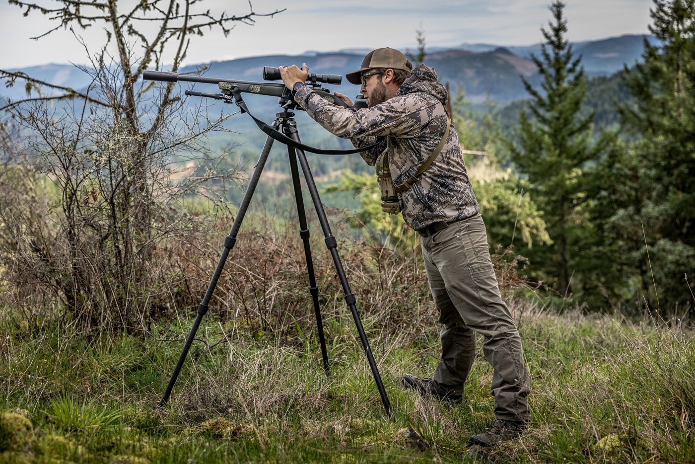 The image shows the author standing behind a tripod extended to chest height, his rifle mounted to the head and his offhand pressing down on the scope. Tripod standing is one of the hunting shooting positions built for seeing over obstacles, letting a hunter clear grass and brush that would block a seated shot. The position is well suited to coyote, hog, and antelope hunting on flatter terrain, though it takes longer to set up and is less stable than sitting. He spreads his feet shoulder-width apart and leans slightly into the tripod, loading it with forward pressure to steady the rifle. A key technique here is isolating the shoulder, applying roughly 60% of the pressure with the dominant hand and about 40% with the shoulder. That approach reduces the wobble zone and limits the effect of heartbeat and breathing on the sight picture. The author still keeps light shoulder contact on the buttstock, just without driving into it, a skill that rewards practice at the range.