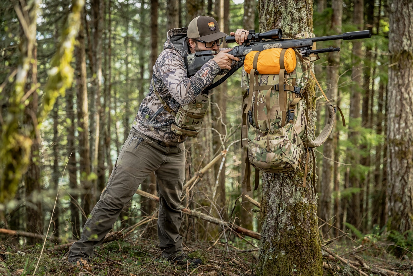 The photo shows the author standing at a tree with his frame pack flipped upside down and lashed to the trunk by its hip belt, the rifle resting on the bottom of the pack at chest height. This is the frame pack standing position, one of the more unorthodox hunting shooting positions but a stable way to take a standing shot in the timber. Most hunters end up bracing against a tree at some point, and securing the pack as a shelf is far steadier than simply leaning on the trunk. The setup works especially well for elk and deer hunting in pine forests, where suitable trees are easy to find. He places the rifle on the pack and either pinches the gun to it or presses down on the scope to lock the hold. The main drawback is time, since cinching the pack in place takes longer than other positions and may not suit an alert or moving animal. More than anything, it shows how existing gear can be used creatively to build a solid rest in the field.