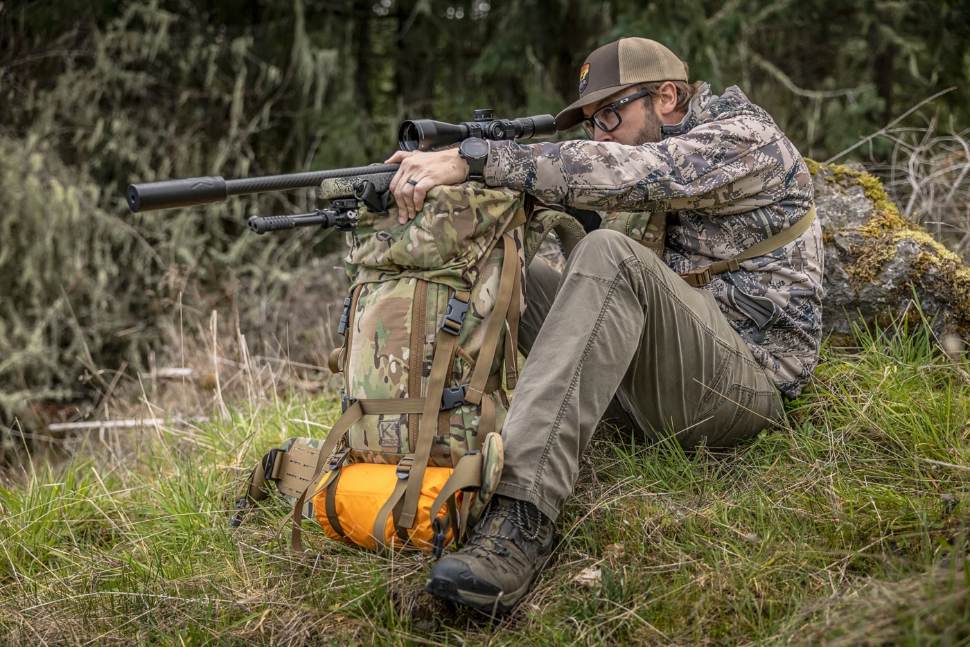 The image shows the author seated on an incline, steadying his hunting rifle across an inverted backpack frame while keeping his weight low against the slope. Among practical hunting shooting positions, the frame pack rest is a go-to for hunters covering long miles with minimal gear. Resting the rifle on the pack frame creates a forward support point similar to shooting off a bag or barricade. Driving the dominant elbow into a bent knee adds a second contact point and shrinks the wobble zone. This seated field position works especially well when a shooter can settle against a tree, boulder, or grade for extra bracing. While less rigid than a tripod, it remains a reliable platform for the kind of shots most hunters actually take afield.