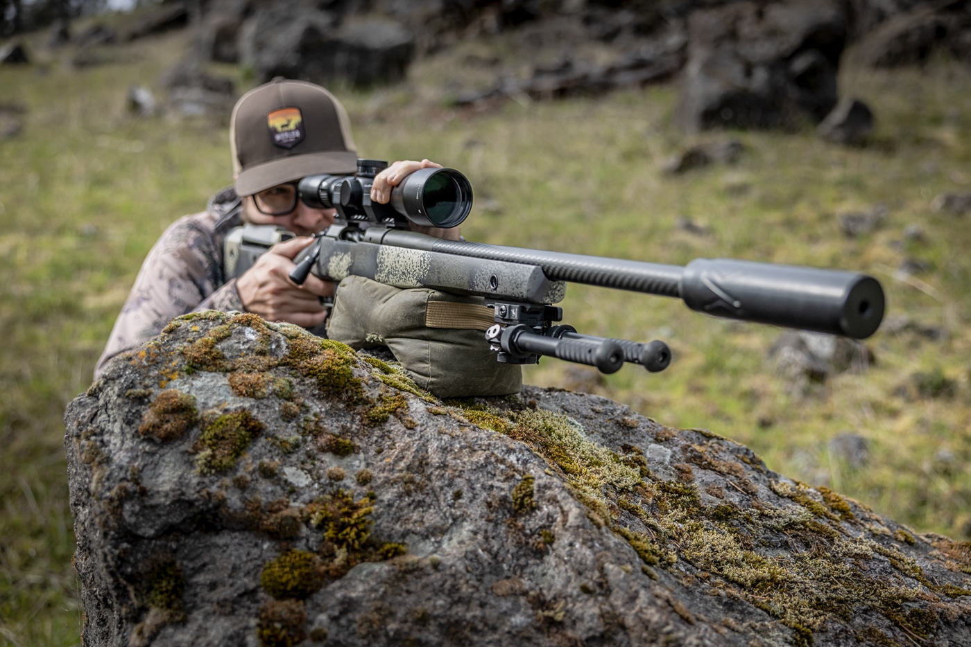 A hunter rests his rifle on a shooting bag that sits atop a large rock, his hands steadying the gun as he settles into the improvised position. The shooting bag is one of the most adaptable tools among field-expedient hunting shooting positions, capable of turning rocks, stumps, fence posts, and fallen trees into stable rests. In this image the boulder provides the elevated base while the bag conforms to its surface and cradles the rifle. Placing the rifle near the center of the gun, just ahead of the magazine, reduces wobble and keeps the hold steady. The shooter's offhand presses down on top of the scope to control movement and recoil. Because body position varies with the object used, a hunter may end up seated, kneeling, or standing behind the bag. Practicing this kind of barricade work pays off when a natural rest is the only support available.