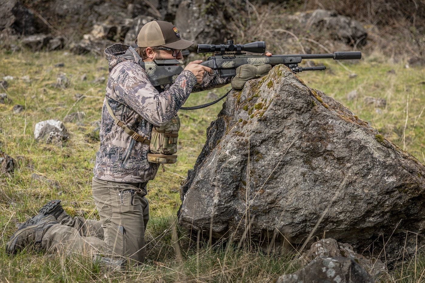 The photograph shows the author kneeling behind a large rock with a shooting bag positioned beneath his rifle to create a firm, level rest. Shooting off a bag is among the most practical field shooting techniques because the bag molds to almost any surface a hunter encounters. On a rounded or narrow object like this boulder, the notched side of the bag faces down so it grips the rock and steadies the platform. Resting the rifle close to its balance point keeps the muzzle from wandering and improves the sight picture. The hunter can pinch the bag and rifle together or push down on the scope to lock everything in place. These small details separate a stable position from a shaky one when the shot finally presents itself.