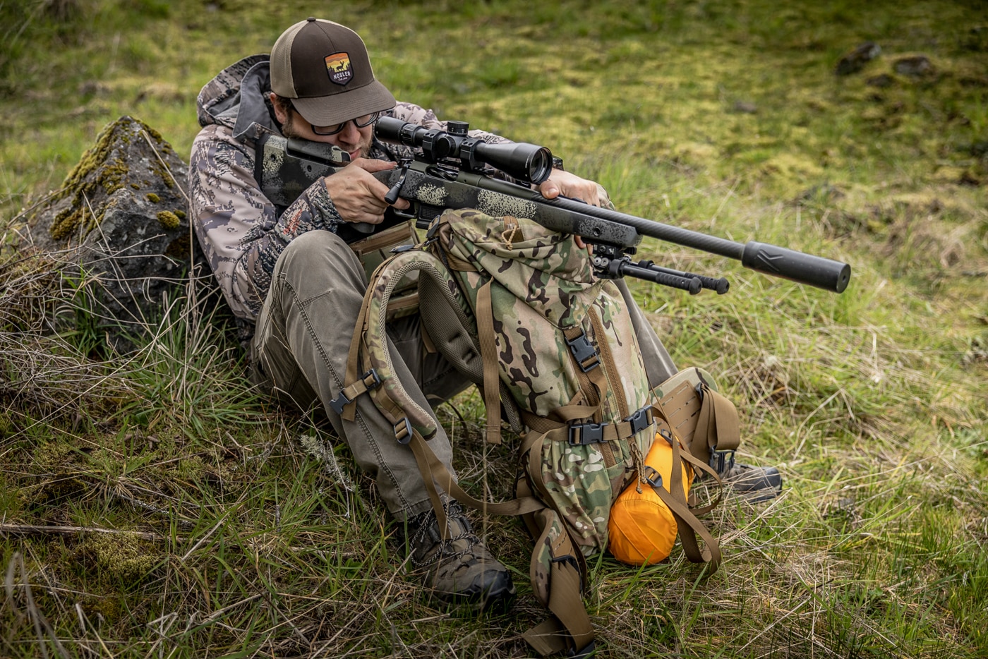 A hunter sits on a grassy slope with his rifle resting on top of an upended frame pack in front of him, his firing-side knee raised to support his elbow. This is the frame pack sitting position, one of several field-expedient hunting shooting positions that turn standard gear into rifle support. Because most backcountry hunters already carry a frame pack, the position adds stability without the weight of a tripod or shooting bag. The seated posture keeps the shooter's center of gravity low, which helps on steep terrain like the slope shown here. The pack frame serves as a forward rest while the raised knee and braced elbow lock the upper body in place. Pulling the rifle firmly into the shoulder, unlike tripod work, helps manage recoil from this support. It is not the steadiest of the field positions, but it offers enough stability for ethical hits out to a few hundred yards.