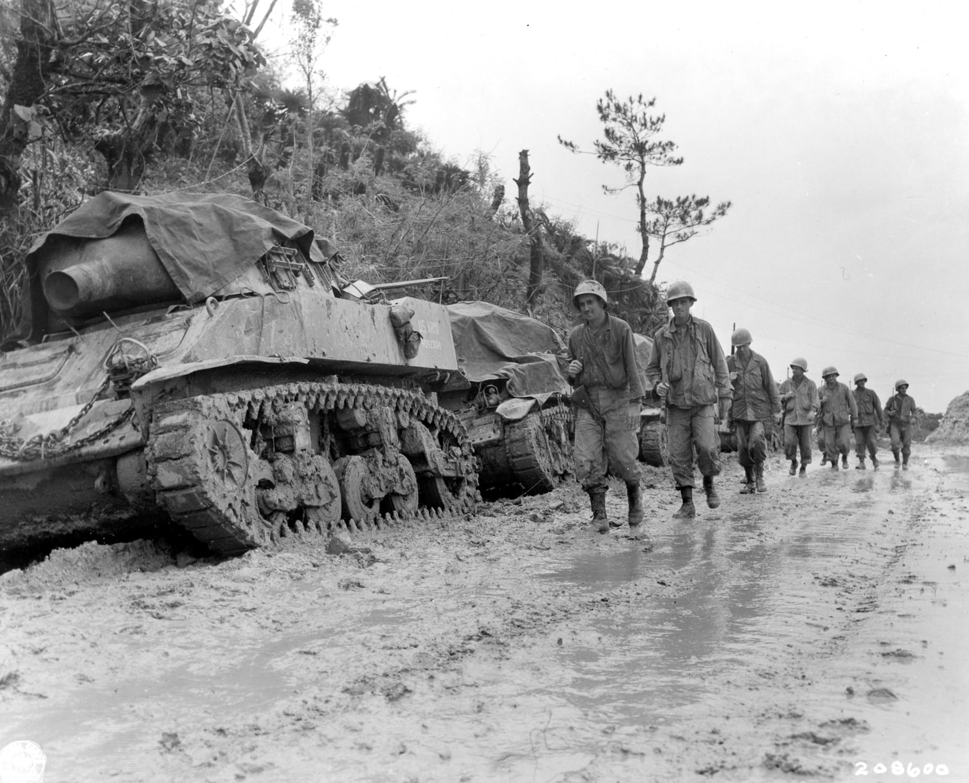 soldiers of 77th Infantry Division march past M8 self propelled howitzer on side of muddy road on Okinawa